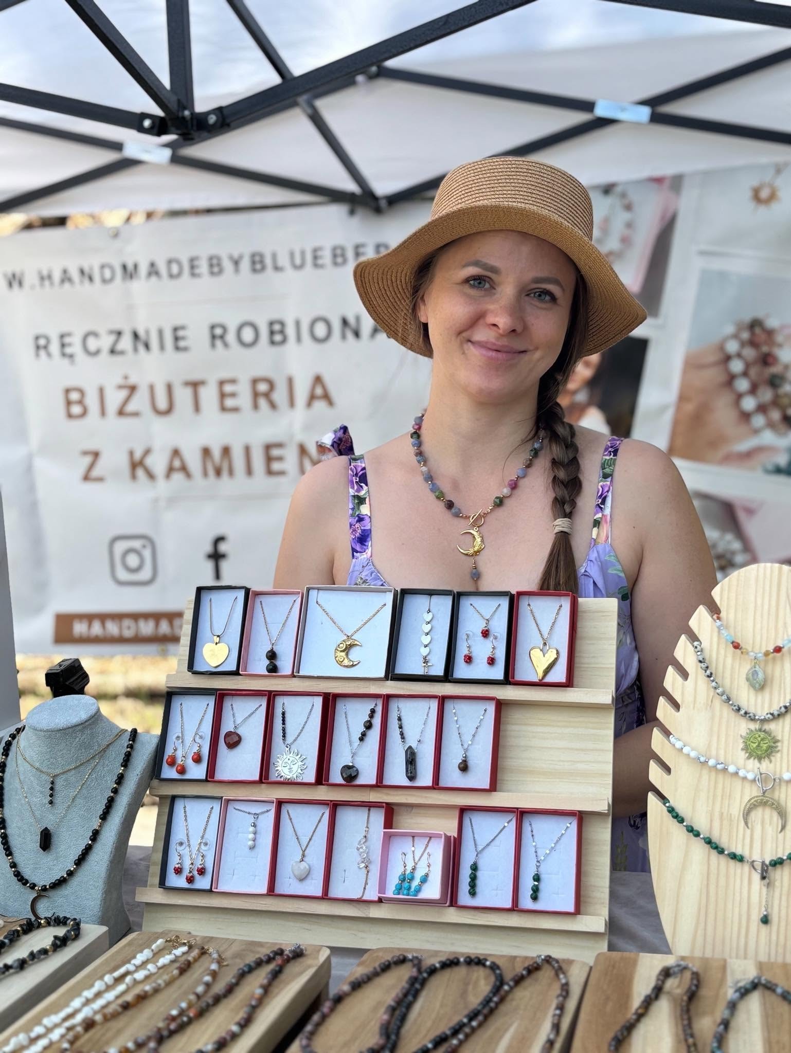 Uśmiechnięta kobieta na targu rękodzieła ze stoiskiem pełnym autorskiej biżuterii z kamieni naturalnych.