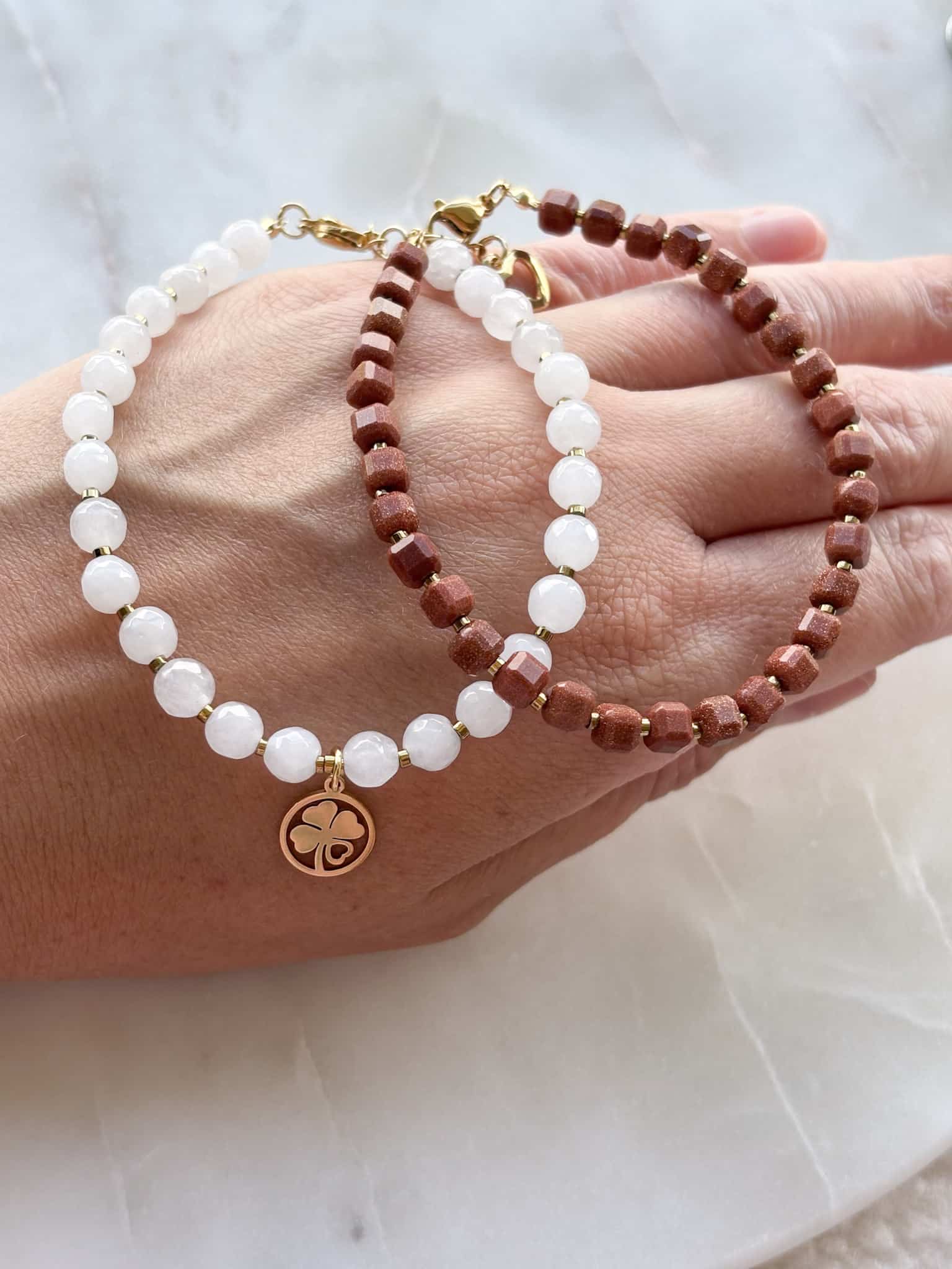 Zestaw delikatnych bransoletek z koralików Zestaw delikatnych bransoletek z koralików – biała z koniczynką i brązowa na dłoni.